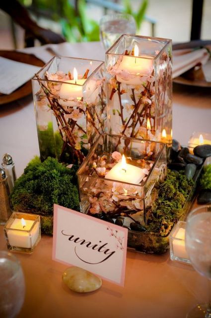 a beautiful wedding centerpiece of moss, pebbles, square glasses with cherry blossom and floating candles, candles around is a romantic idea