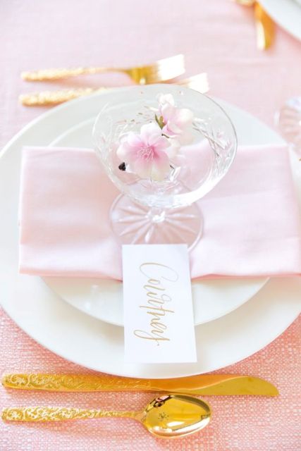 a delicate and glam wedding place setting with neutral plates, a blush napkin and a glass with cherry blossom plus gold cutlery