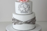 a white wedding cake with silver ribbons, a brooch, silver patterns and monograms plus a pink sugar bloom on top