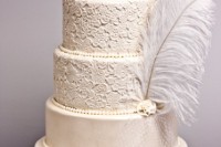 a chic white wedding cake with lace and plain tiers, with beads, a veil and ostrich feathers