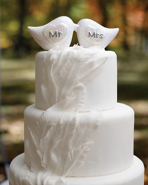 a white plain wedding cake decorated with feathers and with love bird toppers and hearts on them