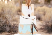 a boho wedding cake in white, blue and gold, with lots of feathers for a strong boho feel