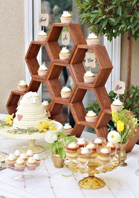 a honey themed wedding dessert table with a honeycomb shelf featuring cupcakes with toppers, greenery and white blooms, a honey cake and some more desserts
