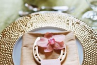 a wedding place setting with a gold charger, a neutral napkin, an orchid and a horseshoe