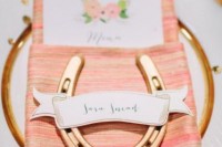 a place setting of a gold rim charger, a colorful coral napkin, a gold horseshoe and a floral card for a rustic wedding