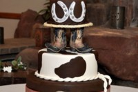 a cowboy wedding cake with a hat, cow print, boots, horseshoes and a romantic couple cake topper