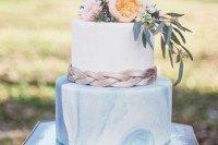 a beautiful wedding cake with a blue marble and a plain white tier, grey braid and fresh blooms and greenery on top