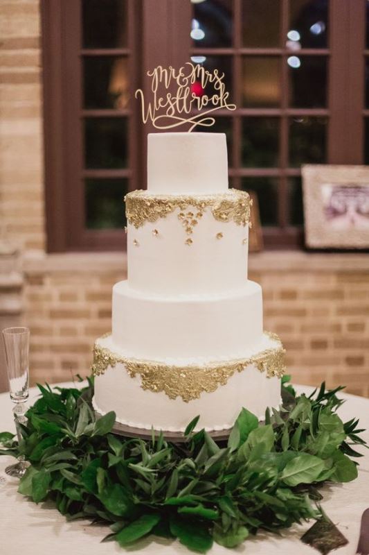 a stylish white wedding cake decorated with gold leaf, with a calligraphy topper and a greenery stand