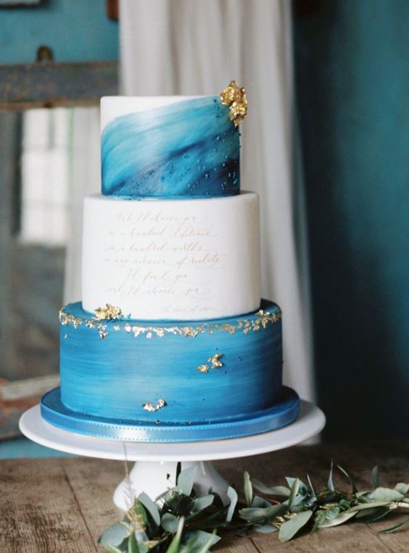 a creative wedding cake with two blue watercolor tiers, a white tier with calligraphy, gold leaf looks very eye-catching