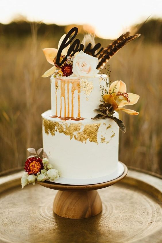 a boho white wedding cake with caramel drip, gold leaf, bold fall blooms and feathers plus a calligraphy topper
