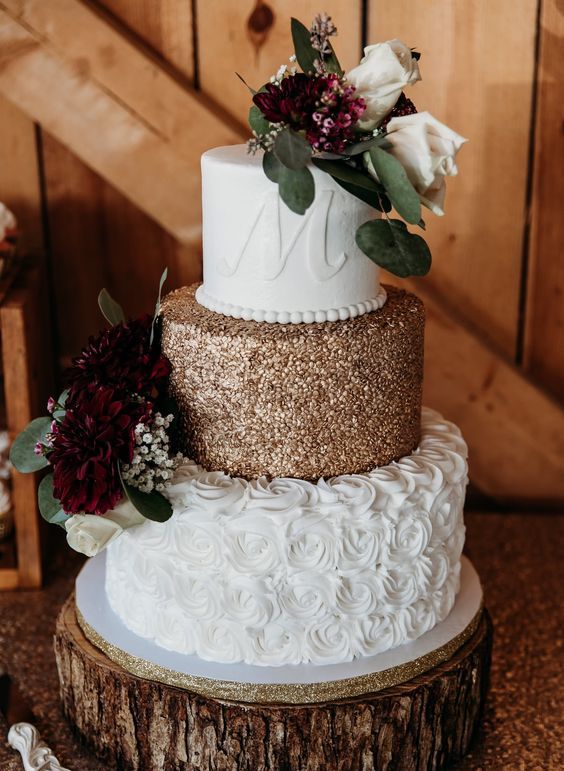 a bold wedding cake with a gold and a white floral tier, with a monogram, white and burgundy blooms and greenery