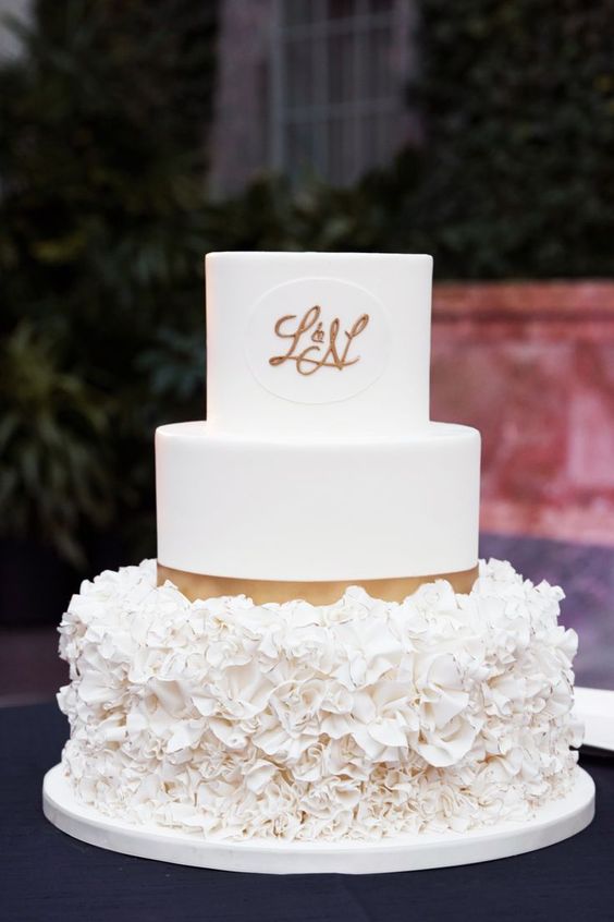 a cute glam wedding cake in white with plain and ruffle tiers, with gold ribbons and gold monograms is a chic idea
