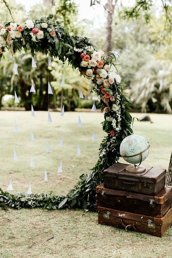a floral and greenery round wedding arch with paper planes, a stack of vintage suitcases and a globe on top
