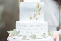 a lovely wedding cake with a white, watercolor blue and calligraphy tier, gold leaf and greenery that covers it