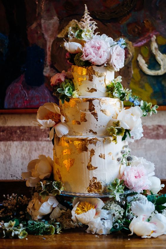 a lush and bold wedding cake with gold leaf, greenery and lots of pink and white blooms looks wow