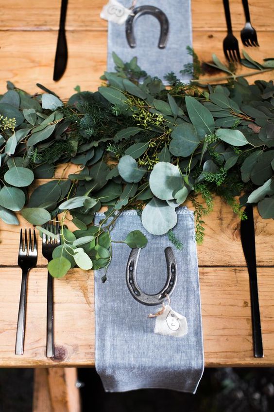 a modern rustic tablescape with a lush greenery runner, simple grey napkins, horseshoes and dark cutlery