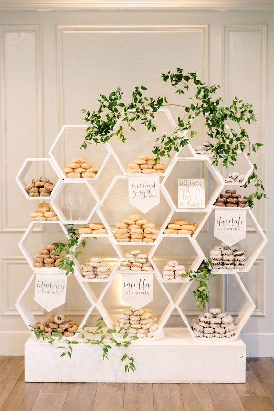 a modern white honeycomb stand with lots of glazed donuts and greenery is a ccreative alternative to a usual wedding dessert table