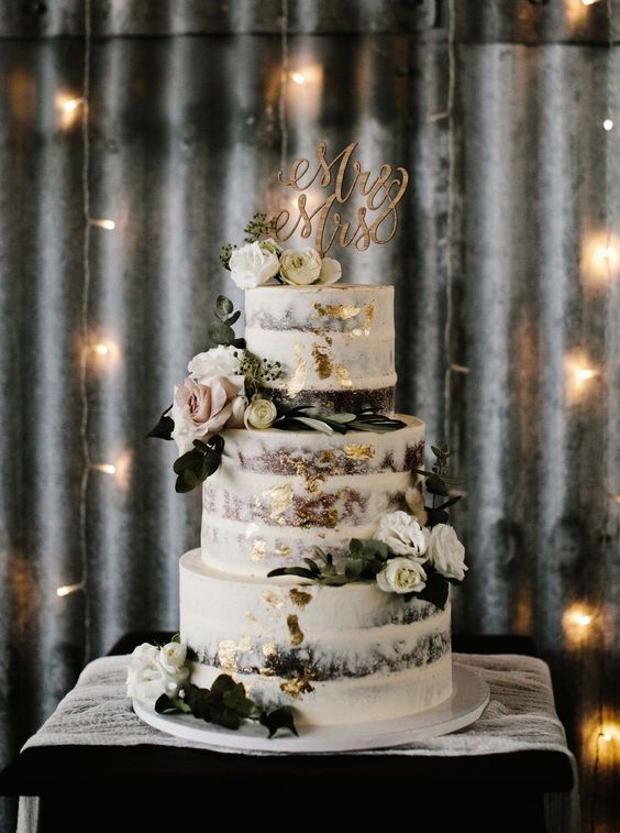 a naked wedding cake with gold leaf, white and blush blooms, leaves and a wooden calligraphy topper