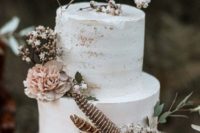a neutral partly naked wedding cake with white blooms, berries, greenery and feathers for a boho or rustic wedding