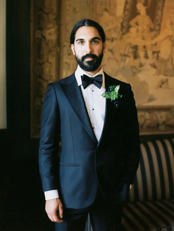 a refined groom's look with a black tux, a black bow tie, a greenery boutonniere, a full beard and a man bun