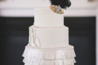a refined white art deco wedding cake with a fringe tier and pearls, a large embellishment and black feathers