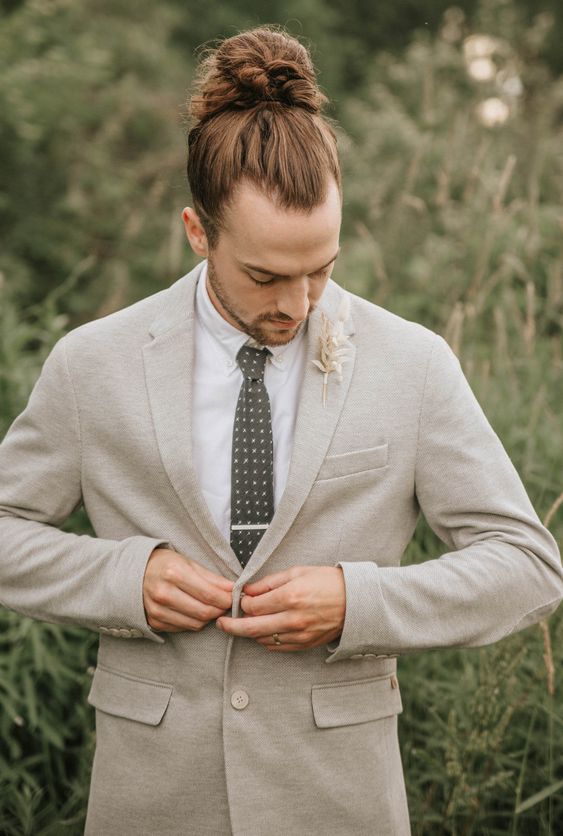 a spring or summer groom outfit with a neutral suit, a printed tie, a dried leaf boutonniere and a man bun on top