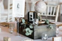 a travel-themed wedding centerpiece of a wooden box, greenery, a candle, a vintage camera, a globe and a sign