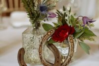 a wedding centerpiece made of vintage glasses, bold blooms and greenery and horseshoes for a rustic wedding
