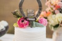 a white buttercream wedding cake topped with pink blooms, greeneyr and a horseshoe with a sign
