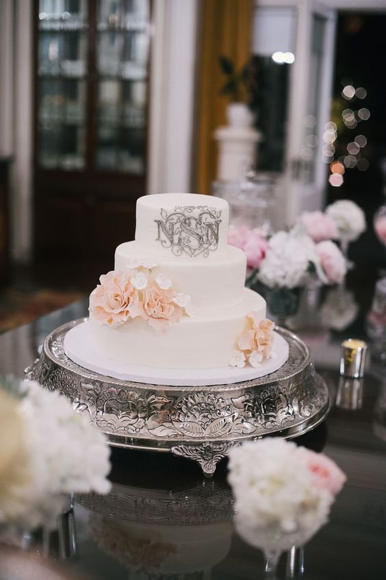 a white wedding cake with white and peachy sugar blooms and a silver patterned monogram for a glam wedding