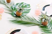 a bright tropical tablescape with a leaf table runner, pineapples, gold cutlery and black chargers