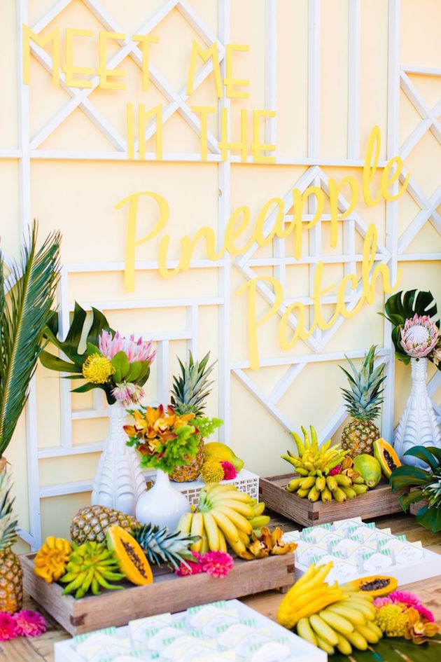 a tropical fruit table decorated with tropical leaves with a yellow neon sign over it