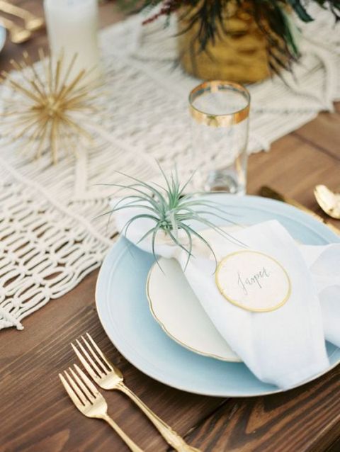 a boho wedding tablescape with a woven table runner, a blue placemat and neutral plates, an air plant as an accent and a favor, some gold touches