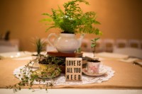 a chic wedding centerpiece of a doily, greenery in vintage teacups, a stack of books and a teapot with ferns growing