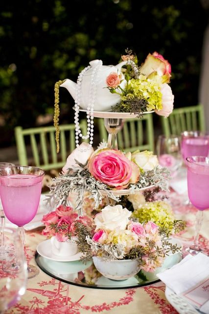 an eye-catchy wedding centerpiece of a mirror, a couple of stands, bright blooms, greenery, pearls and a teapot on top