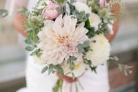 a neutral and pastel wedding bouquet with a light pink dahlia, neutral blooms and pink ranunculus, greenery and a bit of thistles is a gorgeous arranegement