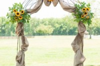 a rustic wedding arch decorated with burlap, greenery and sunflowers looks cool and very cozy