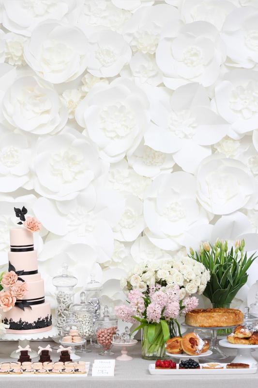 a Parisian bridal shower sweets table with a black and pink cake, pink and white blooms, various candies and pies plus a white paper flower wall