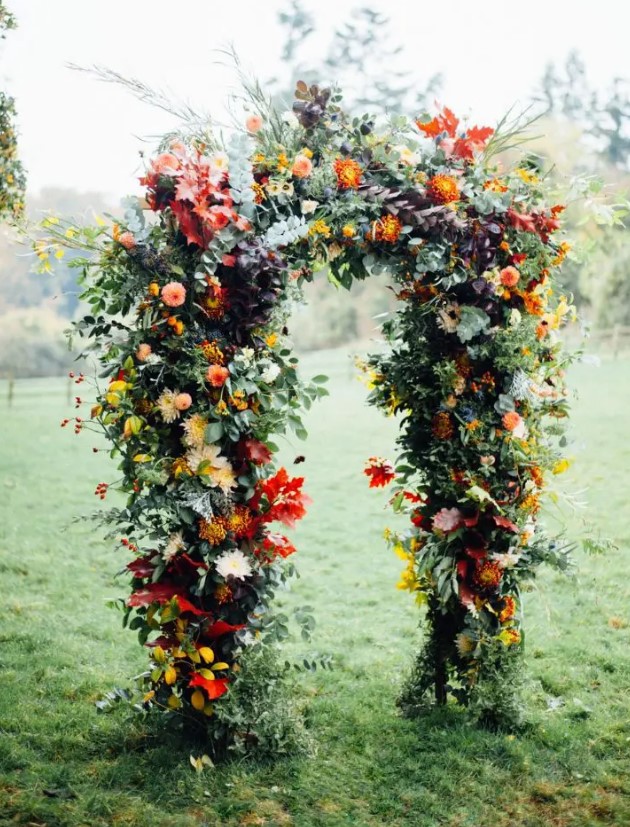 a beautiful and colorful rustic fall wedding arch with greenery, red, yellow, pink blooms, including dahlias and mums, colorful fall leaves is a lovely idea for the season