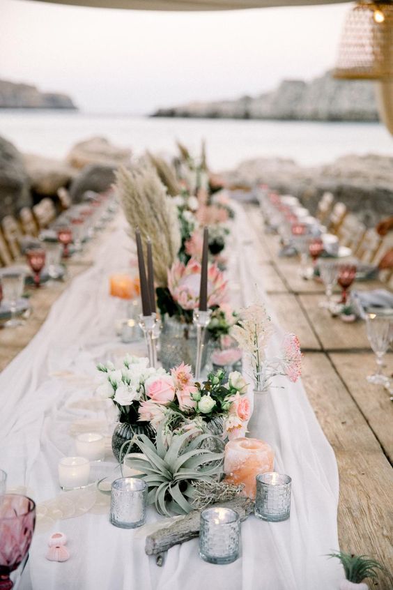 a beautiful wedding table runner of whiet fabric, candles, an air plant, blush and white blooms and greenery, driftwood, tall and thin candles and pampas grass