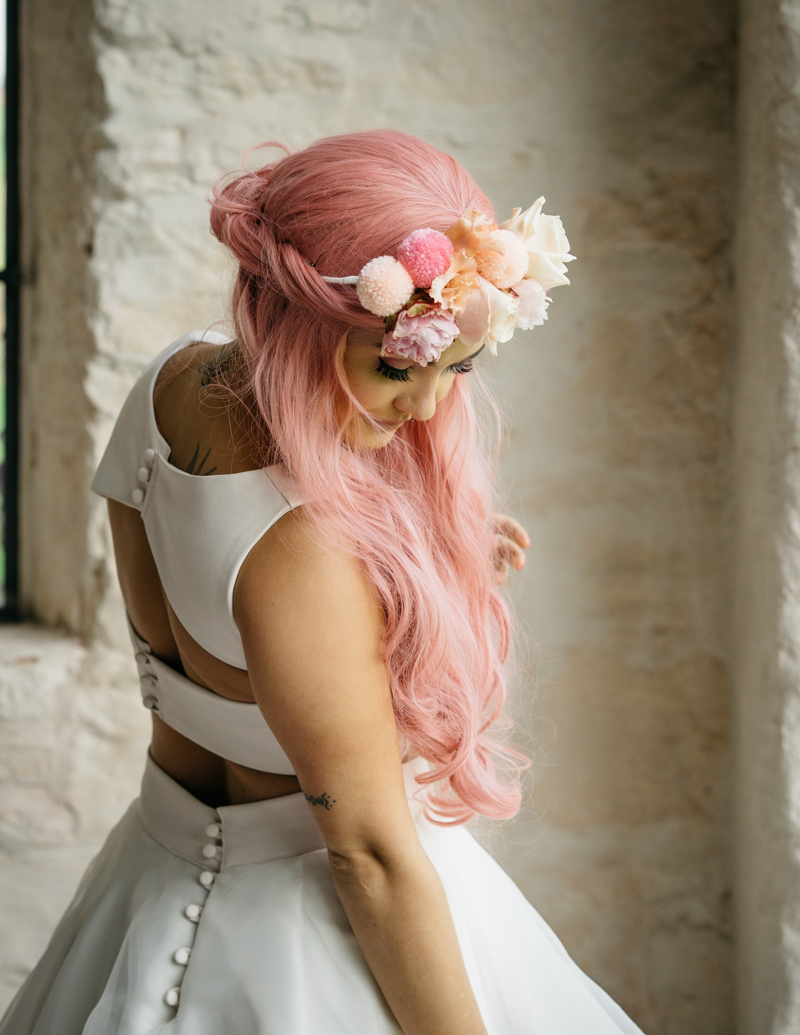 a bride wearing a creative modern wedding dress with cutouts on the back and pink wavy half updo with a floral and pompom crown