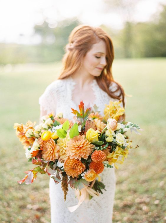 a bright fall wedding bouquet of orange and yellow dahlias, pink and yellow ranunculus and roses, greenery and fall leaves