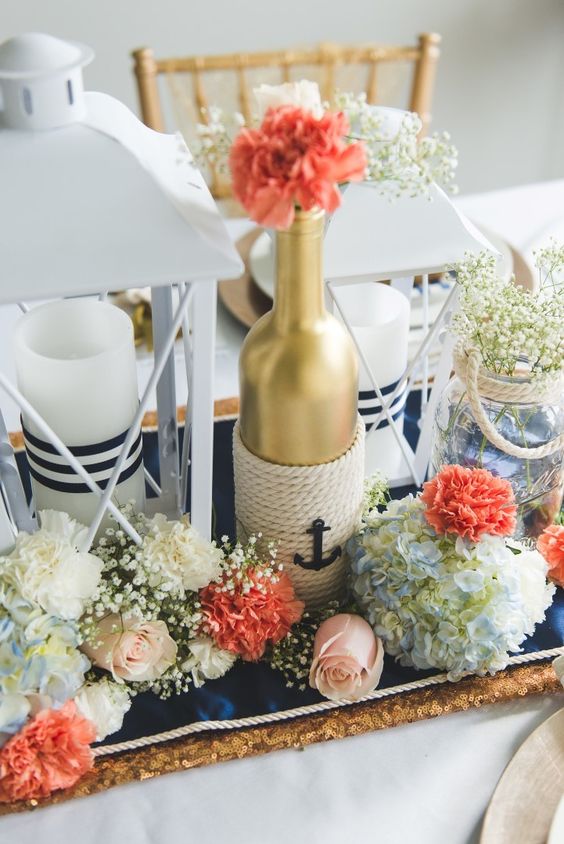 a chic centerpiece with white, coral and blue blooms, a gold bottle with rope, a candle lantern and a jar with baby's breath