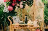a chic rattan tropical bridal shower cart with lots of refreshing drinks, fruits, blooms and a rattan backdrop with monstera leaves and bright balloons