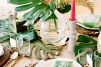 a chic tropcial bridal shower tablescape with wicker chargers, pink candles, monstera leaves and clear glasses looks very modern