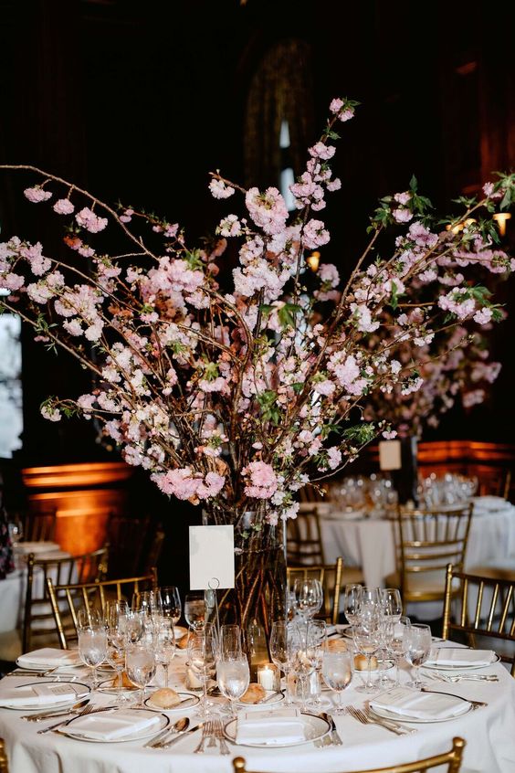 a chic wedding tablescape with neutral linens, tall pink cherry blossom in a vase and gold touches and candles is wow