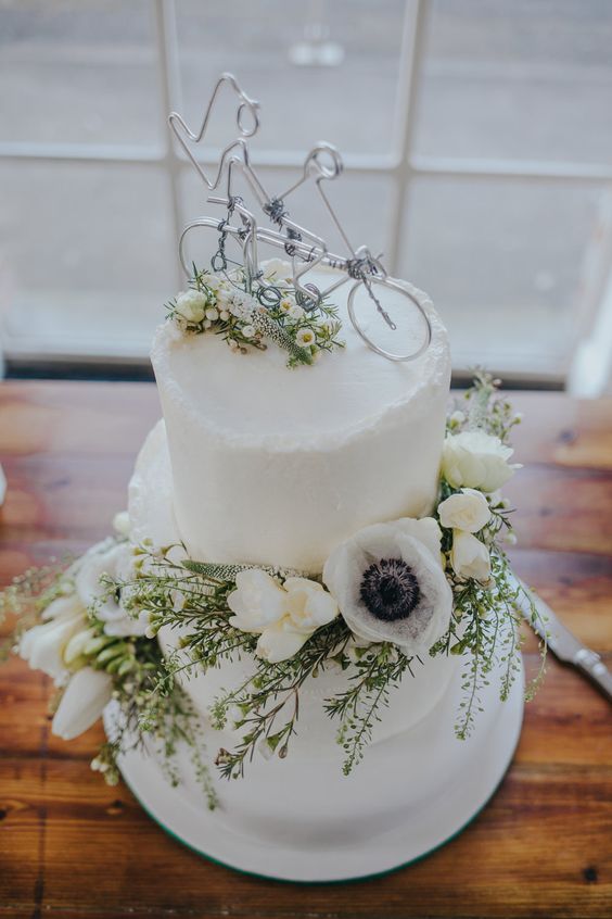 a chic white buttercream wedding cake decorated with white blooms includign anemones and a bike cake topper is wow
