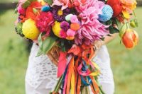 a colorful boho wedding bouquet with red, pink and purple blooms, foliage, colorful pompoms and long colorful ribbons