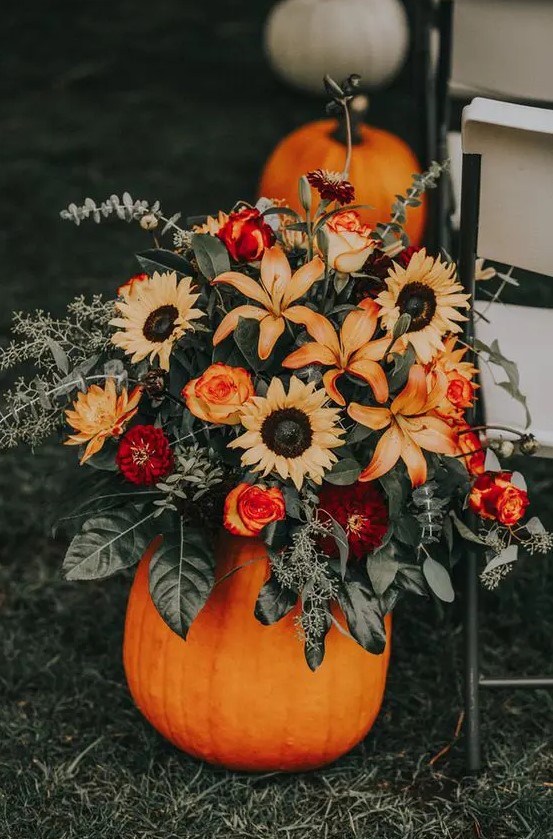 a cool rustic fall wedding decoration of a bold pumpkin, greenery, burgundy, deep red and orange sunflowers, roses and dahlias is a lovely idea for a wedding aisle or table