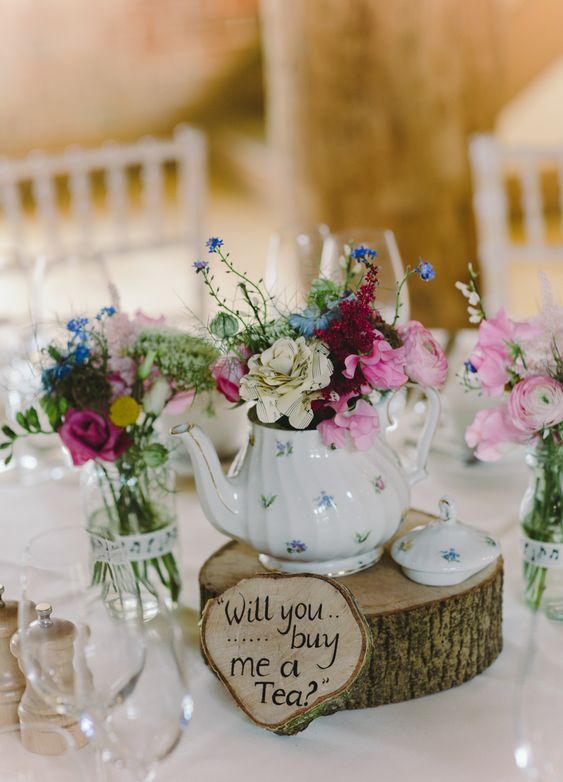 a cool teapot wedding centerpiece of a wood slice, a wood sign, a florla teapot with bright blooms, glasses and bright flowers